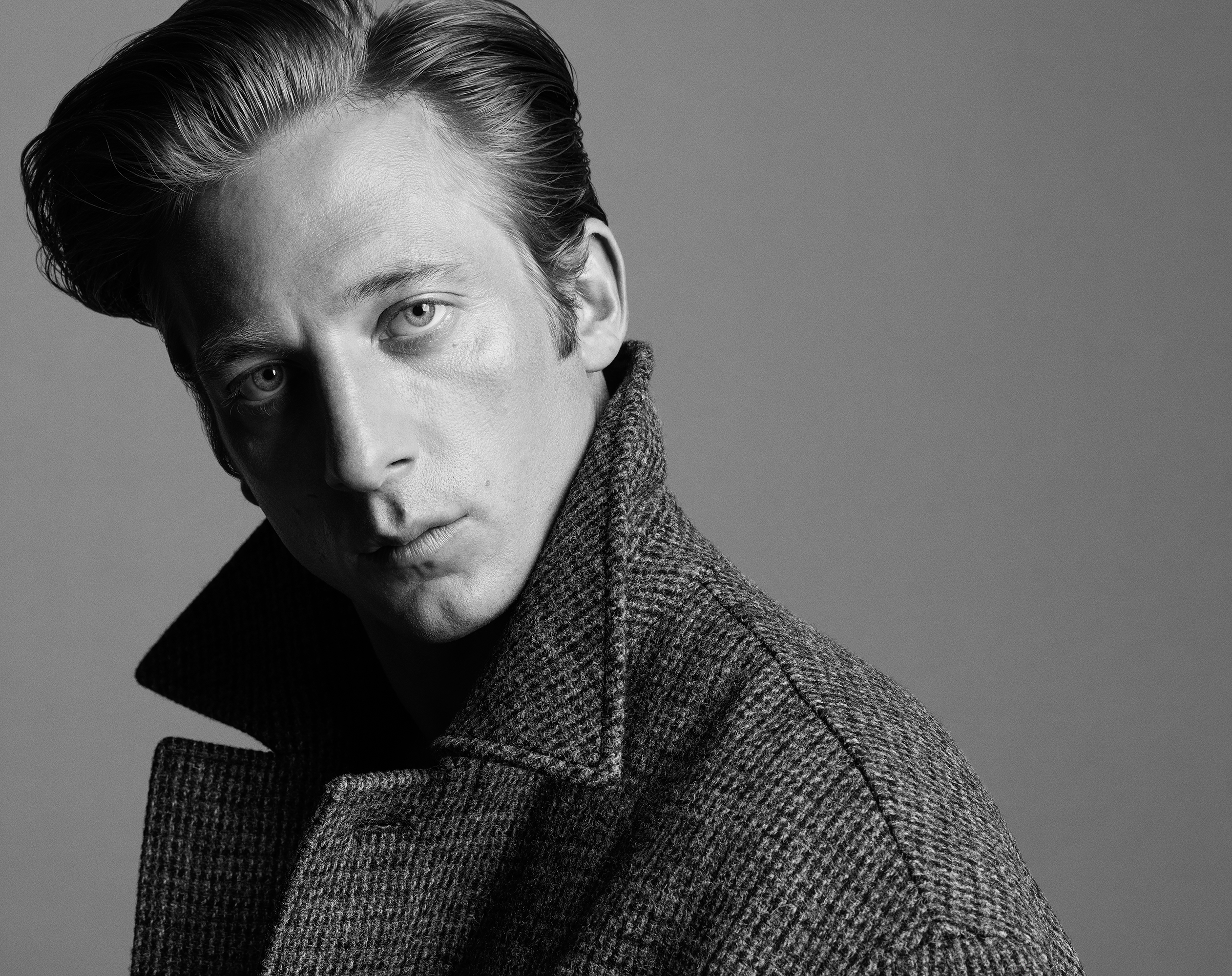 Black and white portrait of a man with dark hair and wearing a tweed jacket with a high collar, gazing intently toward the viewer.