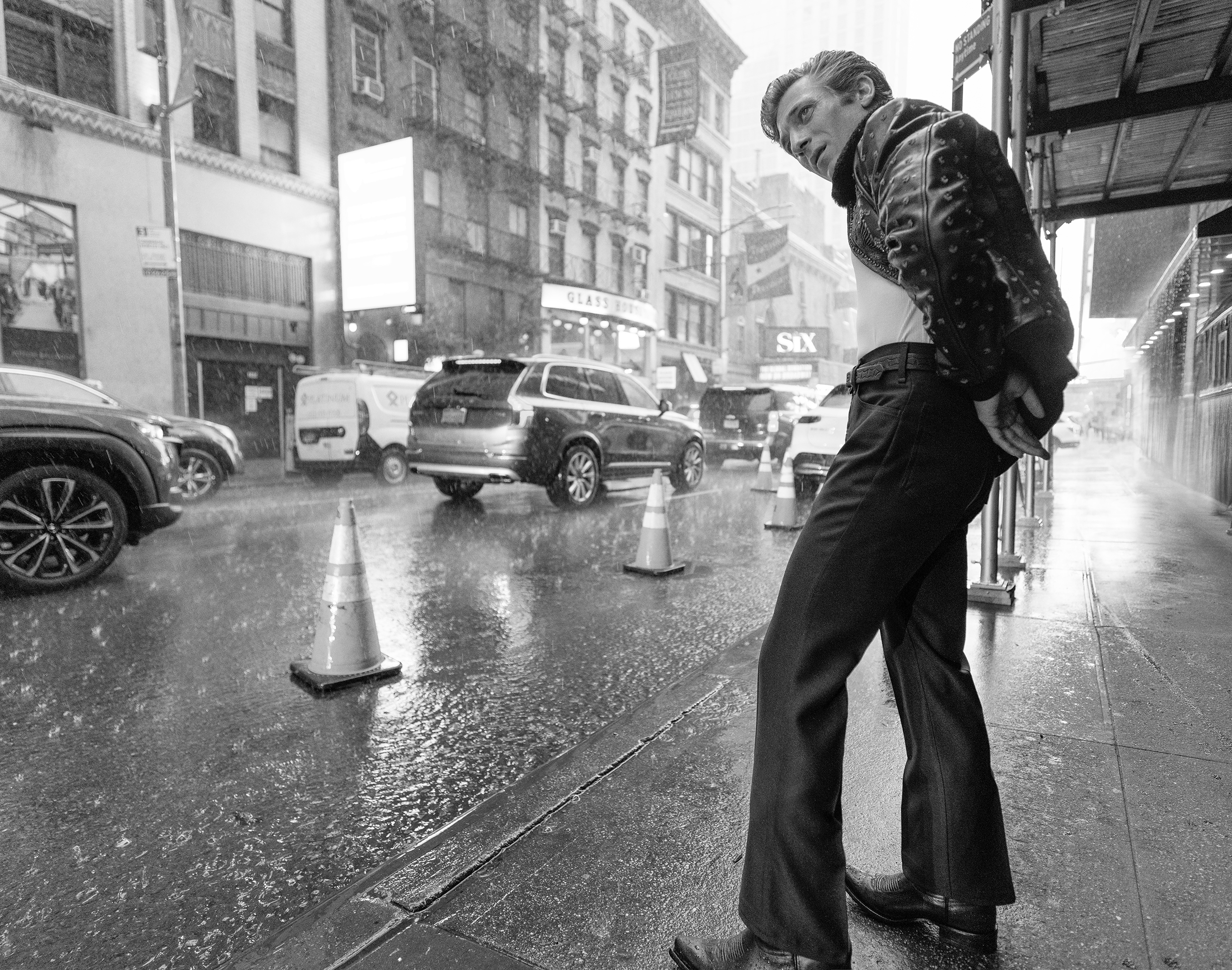 Black and white photograph of a stylish adult male posing outdoors on a rainy city street with traffic and buildings in the background.