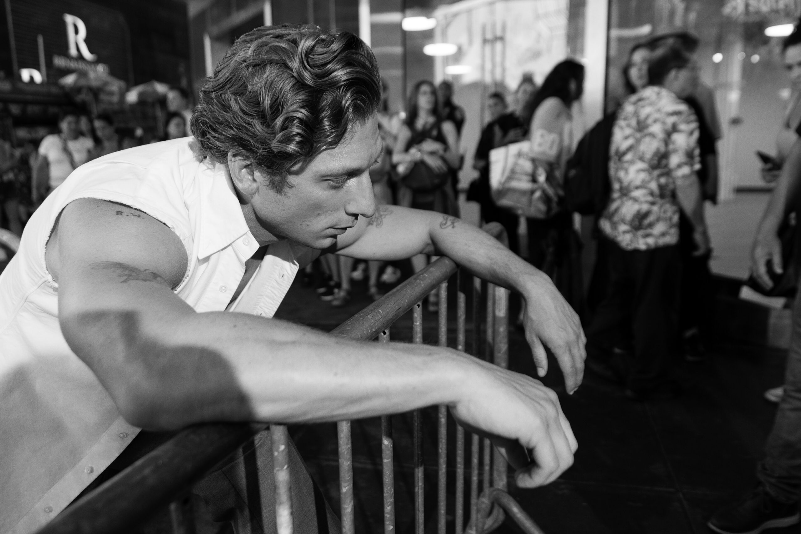 Black and white portrait of a man with dark wavy hair leaning on a metal railing, with a crowd of people in the background.