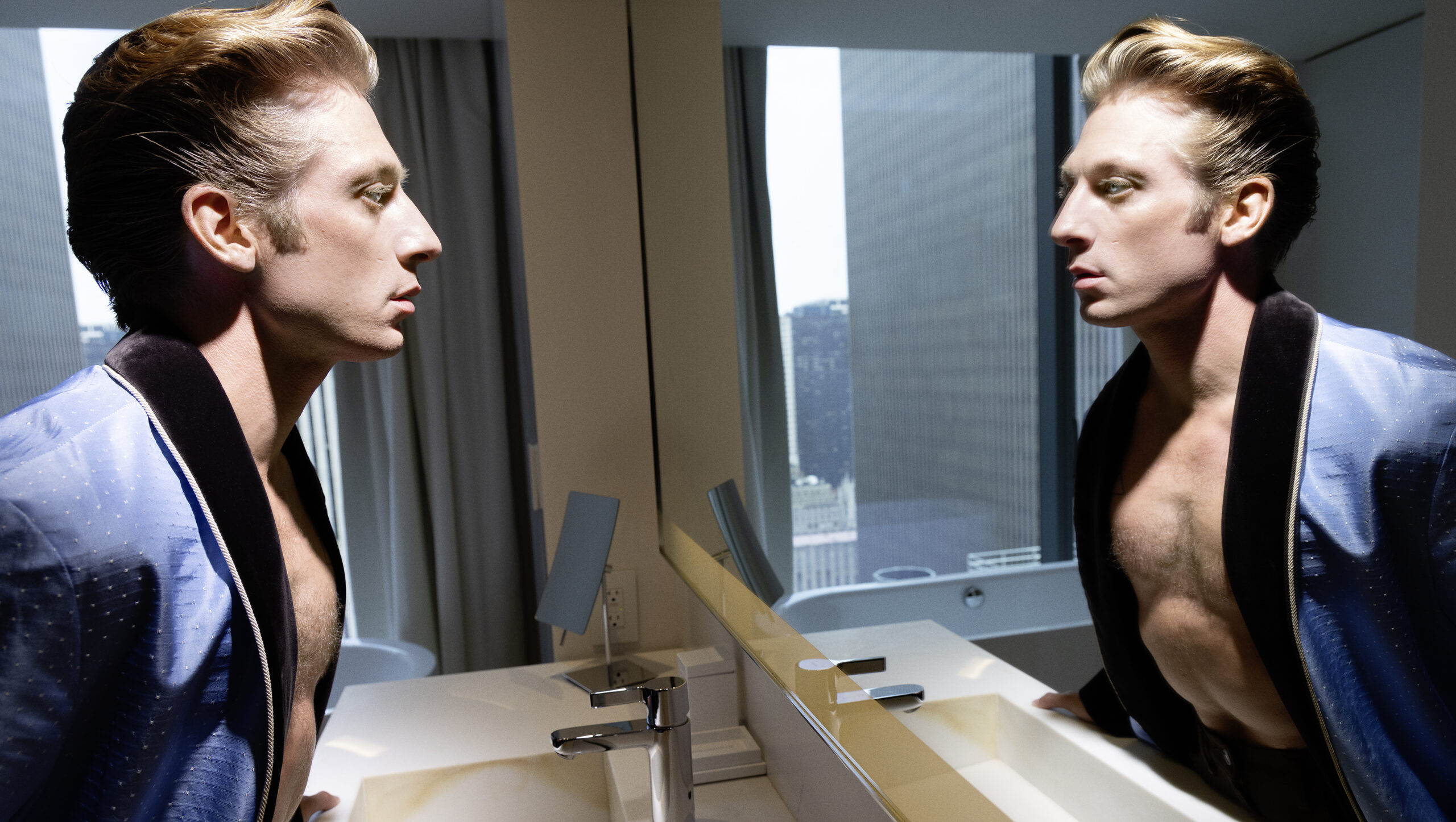 Adult male in blue robe stands in modern bathroom looking in mirror, buildings visible through the window in daytime.