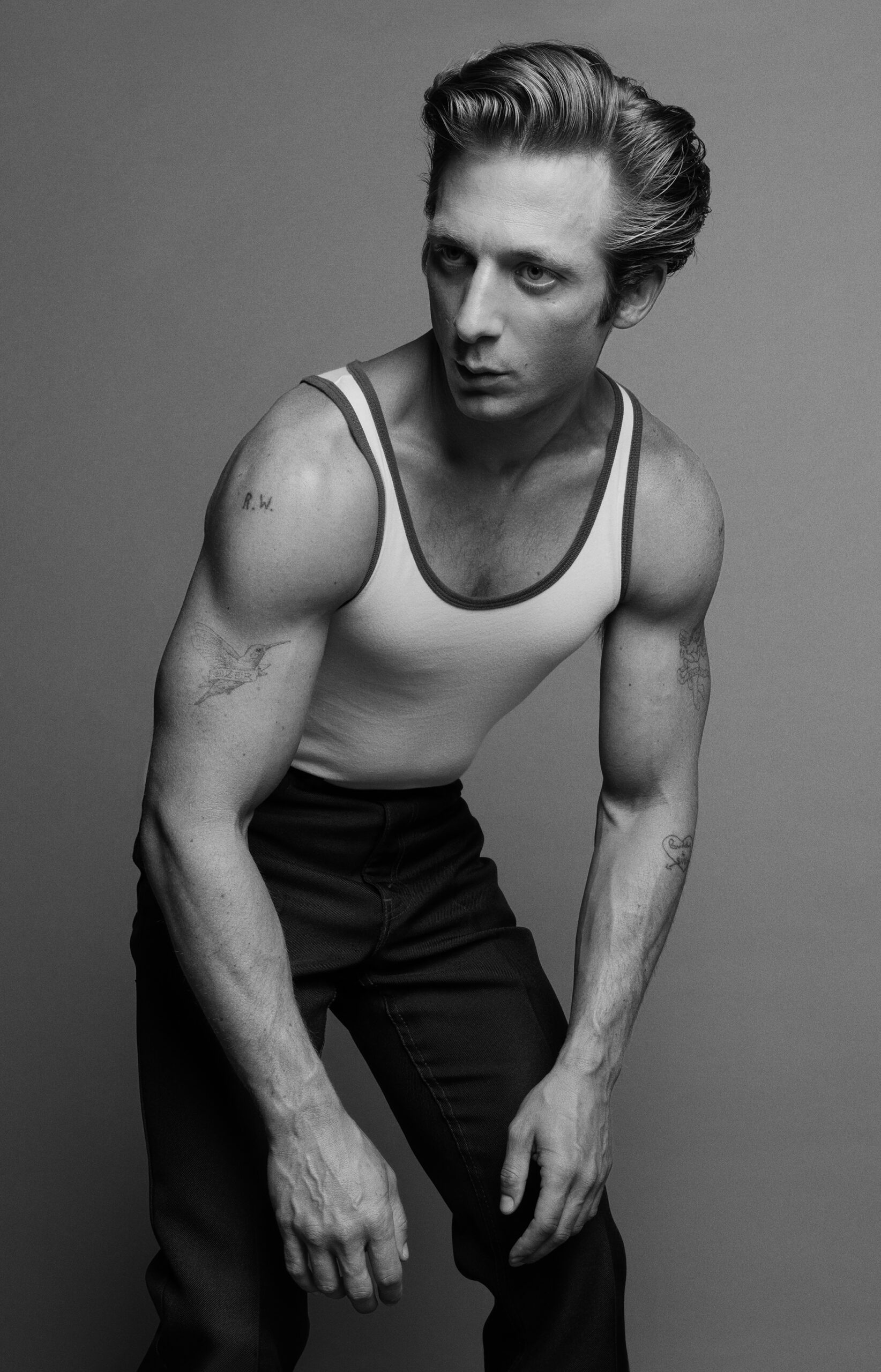 Monochrome portrait of a muscular adult male in a white tank top and dark pants, gazing away against a neutral grey background.