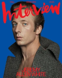 Jeremy Allen White posing in an open grey tweed coat against a vibrant blue studio backdrop for Interview Magazine.