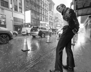 Jeremy Allen White in a dark jacket and trousers standing on a rain-soaked city street, captured in black and white.