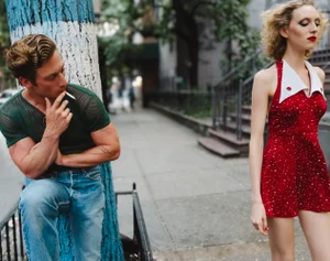 Jeremy Allen White in green mesh shirt smoking a cigarette while a woman in a red dress walks by on a city street.