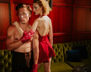 Jeremy Allen White stands shirtless in red studded gloves next to a woman in a backless red sparkling mini dress on a green sofa.