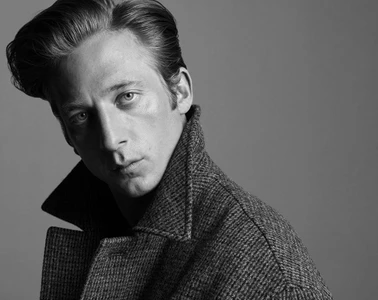 Black and white portrait of Jeremy Allen White wearing a textured tweed coat with an upturned collar against a grey backdrop.