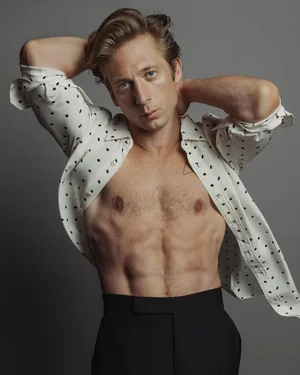 Jeremy Allen White posing in a polka-dot unbuttoned shirt and dark trousers against a grey studio backdrop