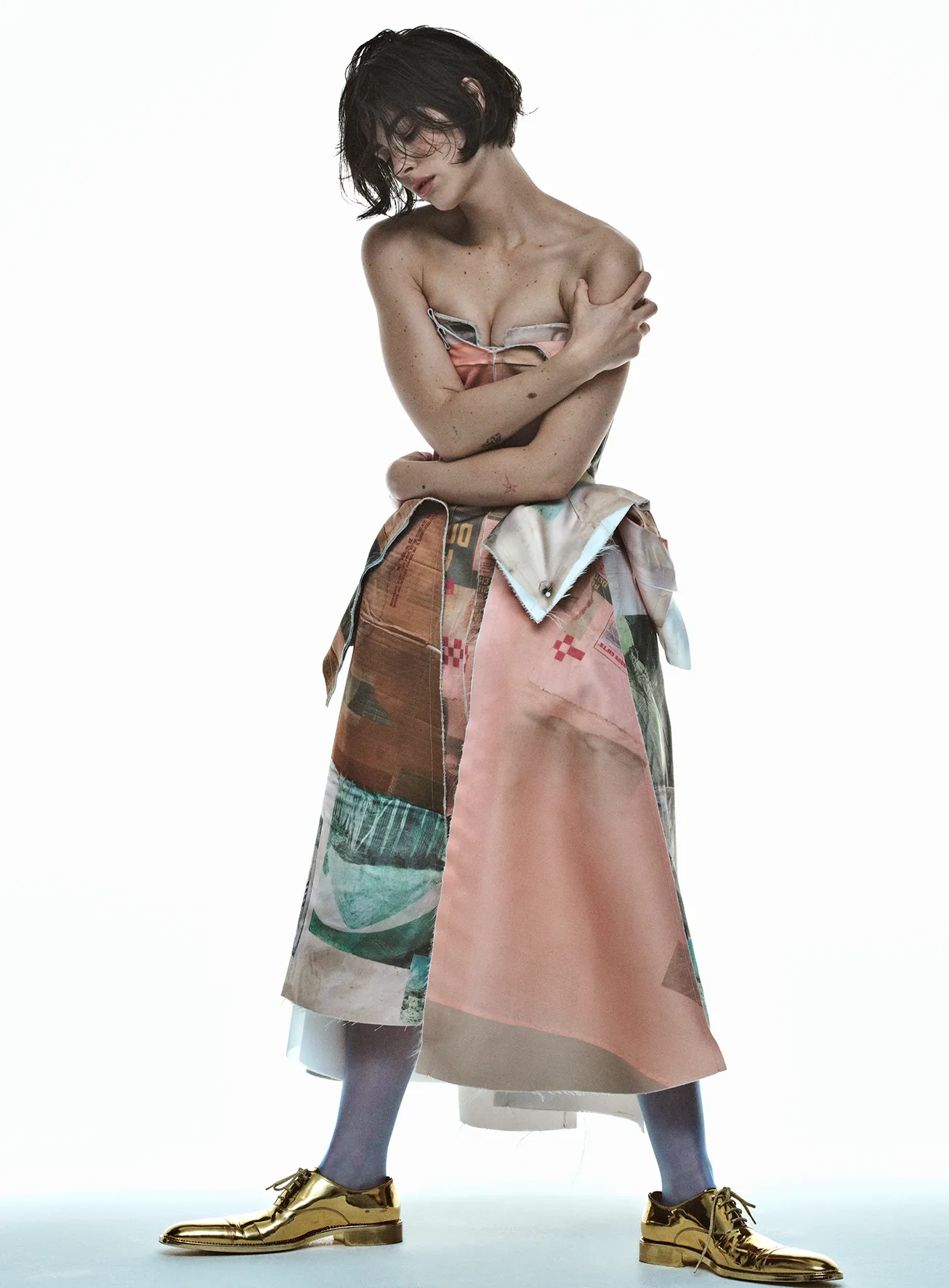 A slender, dark-haired woman poses in a patchwork-patterned strapless dress with golden shoes against a bright white backdrop.