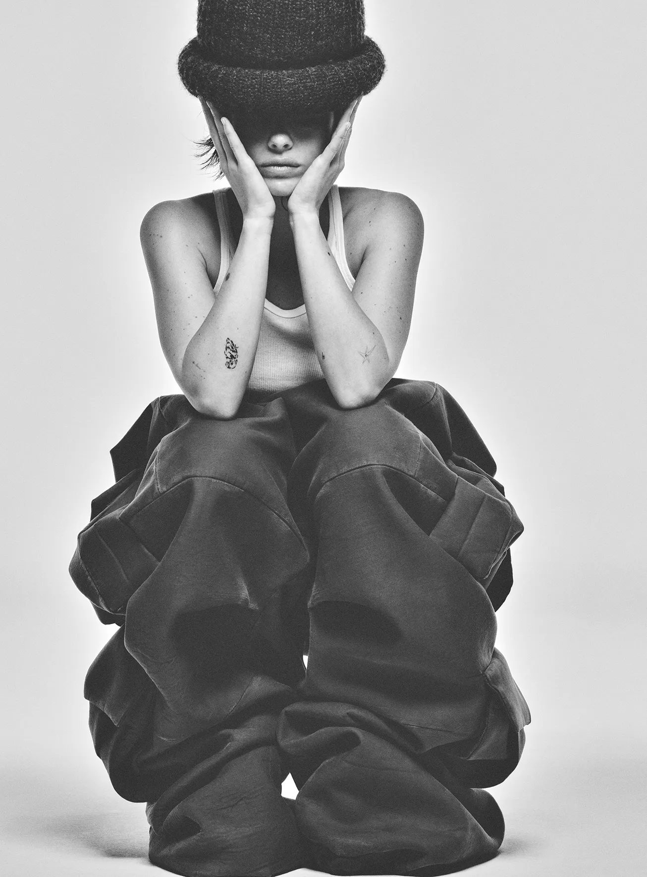 A young woman in a tank top and baggy pants poses in a studio wearing a dark knit hat pulled over her eyes in a black and white shot.