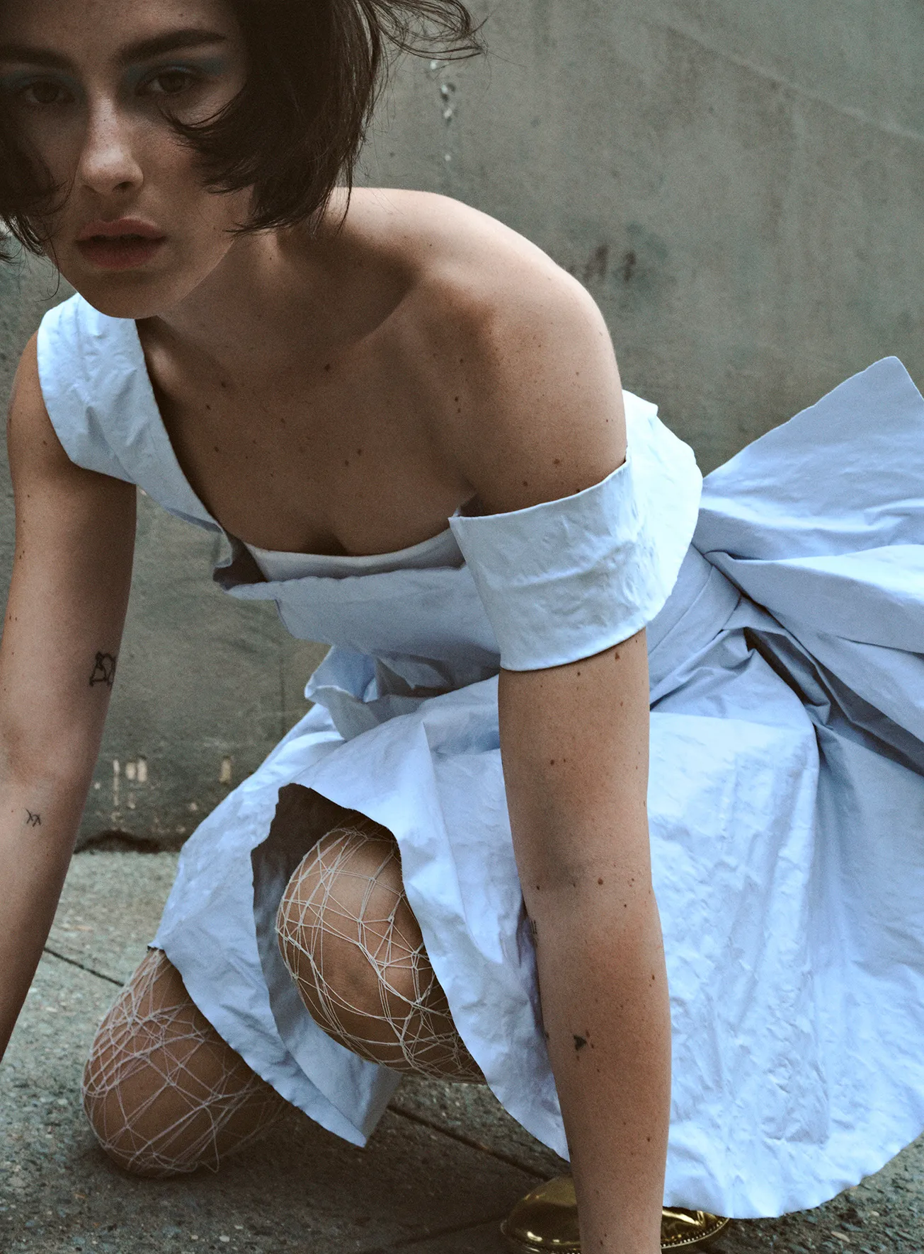Woman in a blue dress poses kneeling on concrete. She wears patterned white tights and gold heels, looking directly at the camera.