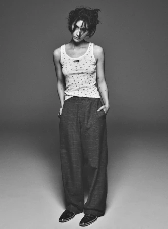 A young adult woman in a patterned tank top and oversized pants, standing indoors against a grey background with a neutral facial expression.