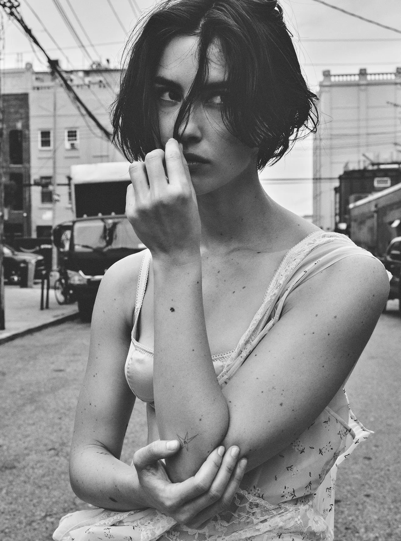 Black and white portrait of a woman with short hair in an urban environment, wearing a lacy tank top, gazing at the camera.