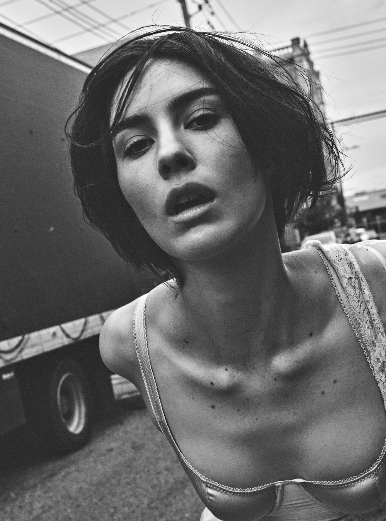 Black and white portrait of a young adult female in a lingerie top outside with an upward angle and truck in the background.