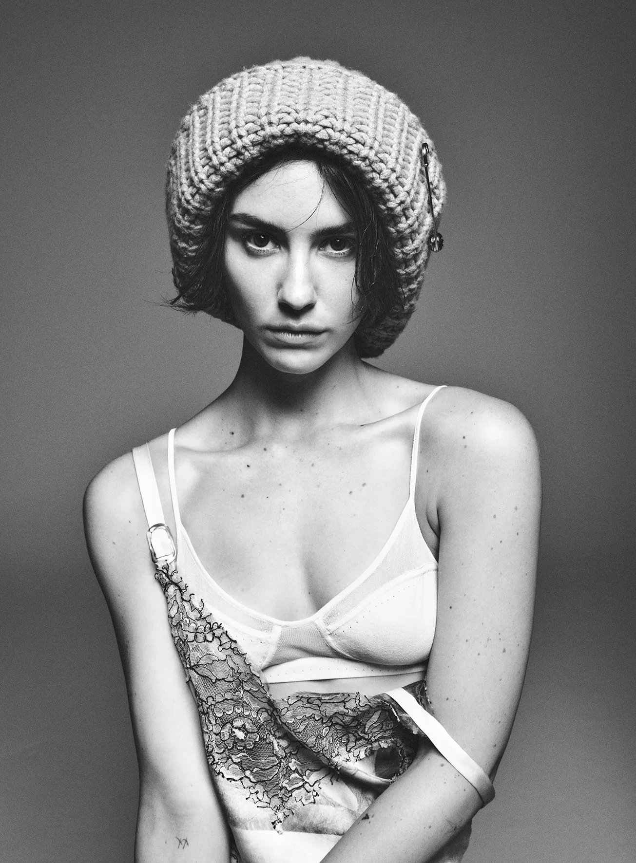 Black and white portrait of a young woman wearing a knit hat, sheer lingerie top, and draped lace fabric, showcasing fashion and beauty.