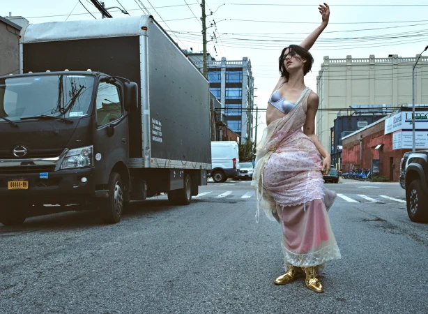 Woman in lingerie and sheer dress poses on an urban street with a truck in the background, showcasing a fashion-forward aesthetic.