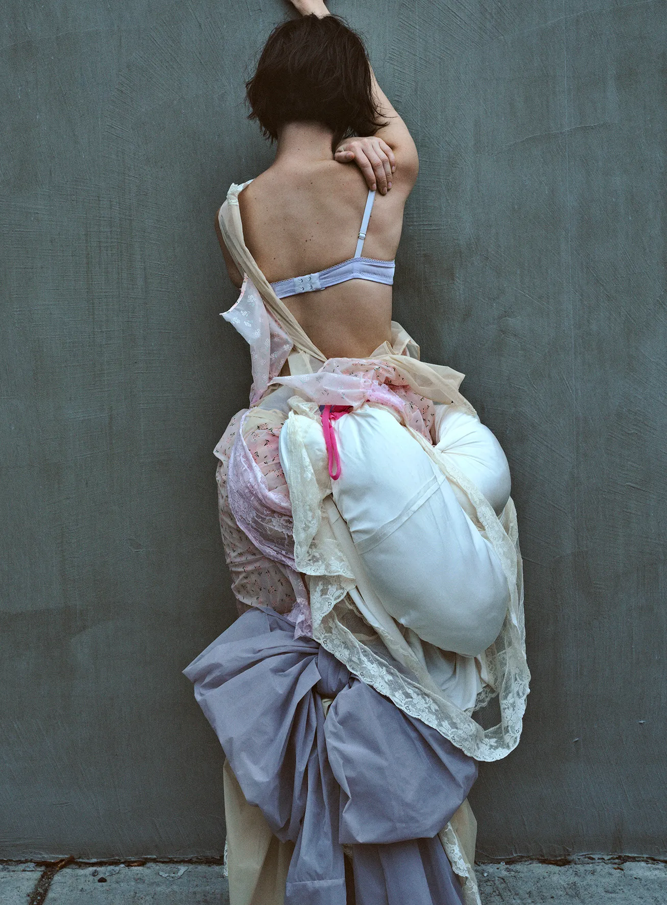 Back view of a person wearing lingerie and an abstract dress made of draped fabric and white objects, leaning against a gray wall.