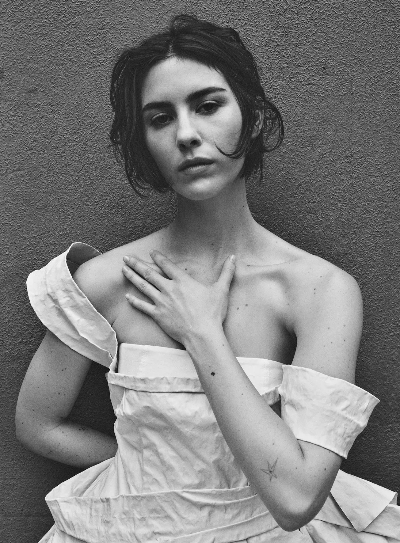 Monochrome portrait of a young woman with short dark hair, wearing a white dress, posing against a simple grey background.