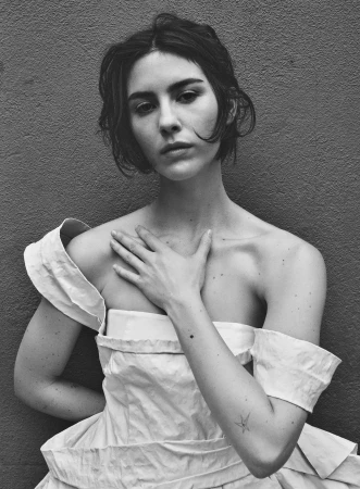 Monochrome portrait of a young woman with short dark hair, wearing a white dress, posing against a simple grey background.