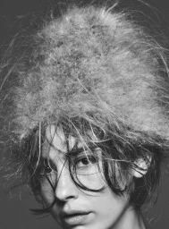 Black and white close-up portrait of Gracie Abrams wearing a large fuzzy textured hat with wispy bangs framing her face.