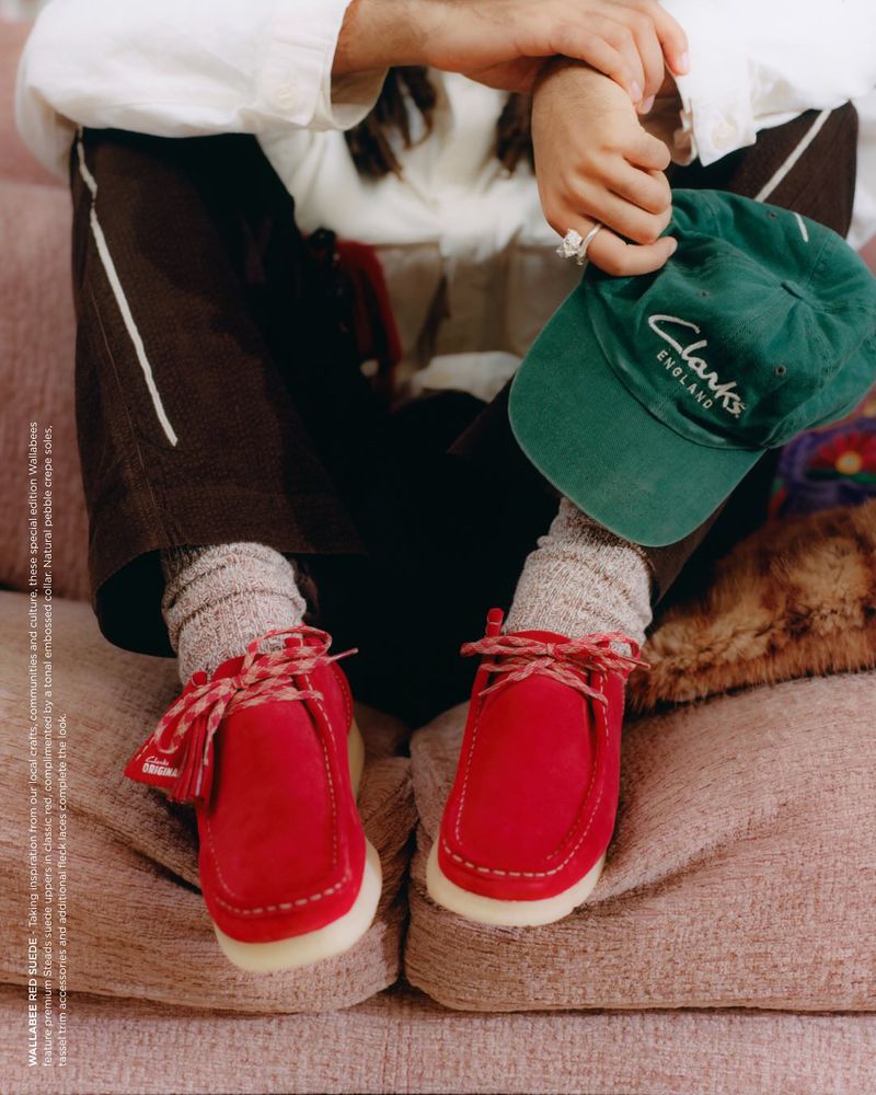 Red suede moccasin shoes with laces, worn with grey socks. An arm, a green hat, and a couch complete the scene.