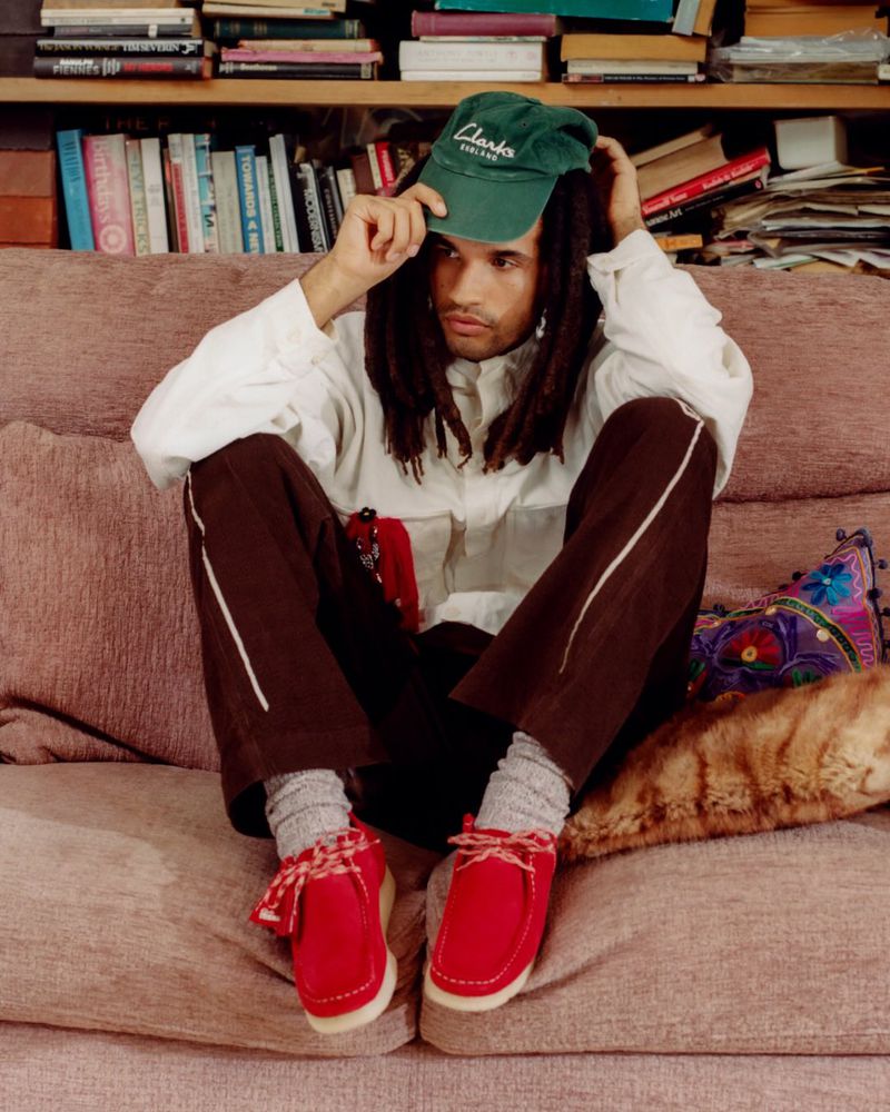 A man with dreadlocks wearing a green cap, white jacket, and brown pants poses while sitting on a couch indoors near a bookshelf.