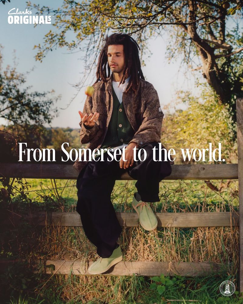 A man with dreadlocks sits on a wooden fence in a rural setting, juggling an apple, wearing Clarks shoes, with the brand logo visible.