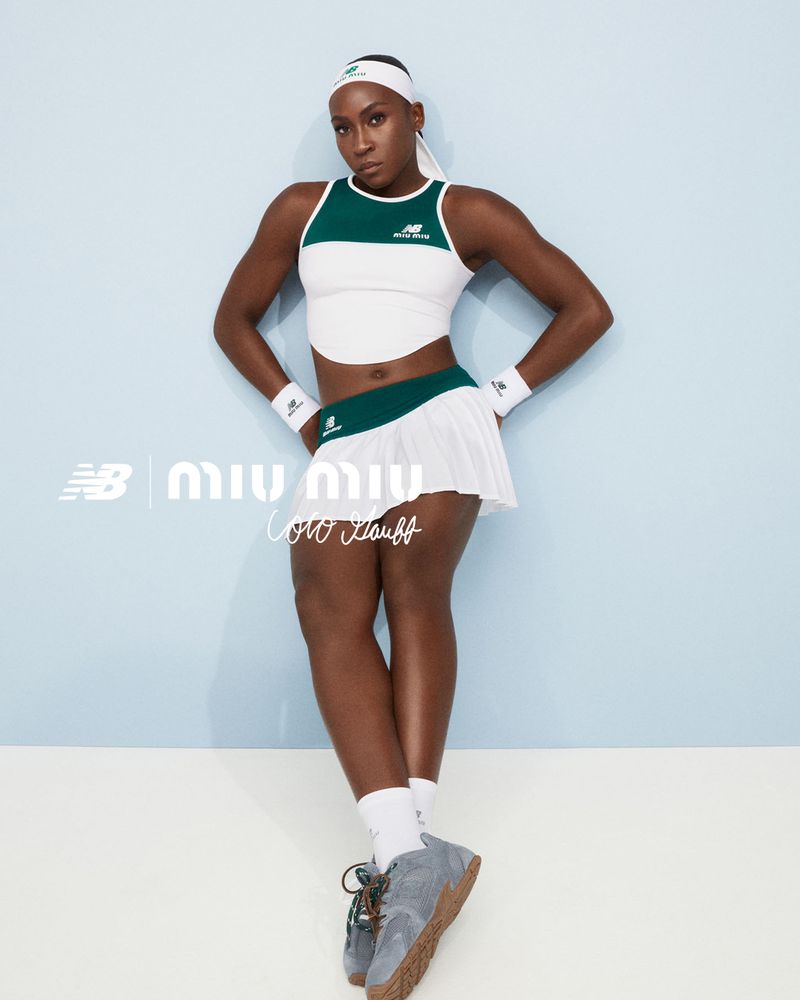 Coco Gauff poses in tennis fashion, wearing a white and green tennis outfit with New Balance grey sneakers against a light blue backdrop.