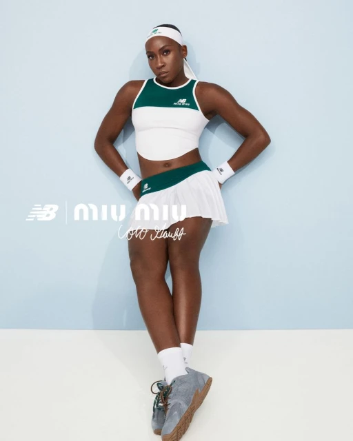Coco Gauff poses in tennis fashion, wearing a white and green tennis outfit with New Balance grey sneakers against a light blue backdrop.