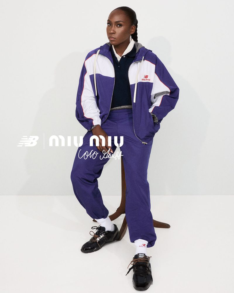 A dark-skinned woman sits on a stool, dressed in a blue and white track suit and black sneakers, posing in a studio setting.