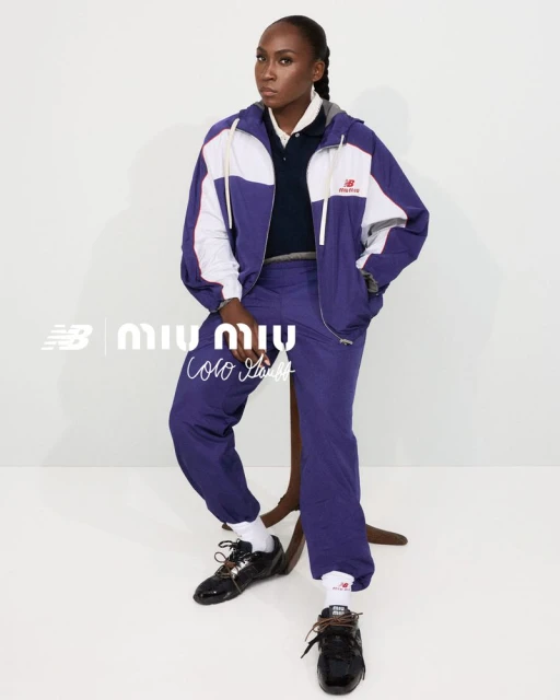 A dark-skinned woman sits on a stool, dressed in a blue and white track suit and black sneakers, posing in a studio setting.