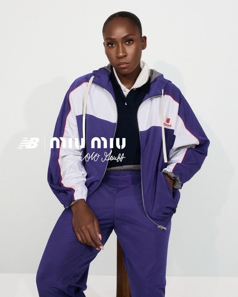 An African American woman poses in a purple and white Miu Miu x New Balance tracksuit in a studio setting with neutral lighting.
