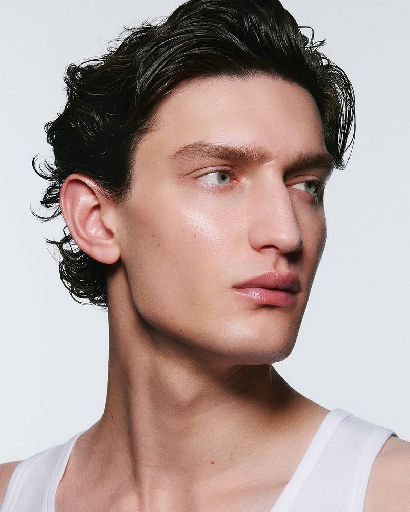 Close-up studio shot of a handsome adult male with slicked-back dark hair, light skin, and a light tank top against a white backdrop.