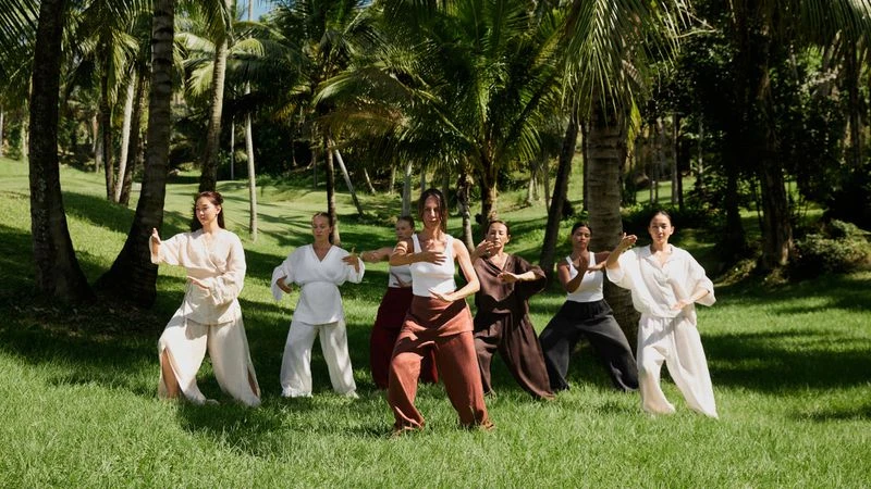 A group of seven women practice Tai Chi in a lush, green outdoor setting with palm trees, wearing casual clothing and light colors in daylight.