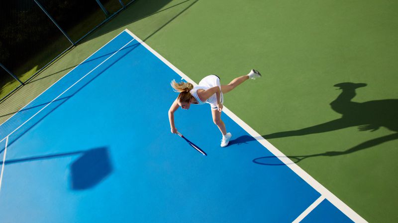 Overhead view of a blonde woman playing tennis on a blue and green court, serving with a racquet in a white tennis dress.
