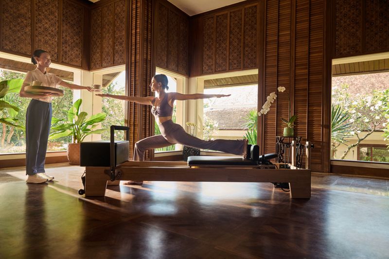 An adult female balances in warrior pose on a Pilates reformer indoors as a server brings her a drink near a window.