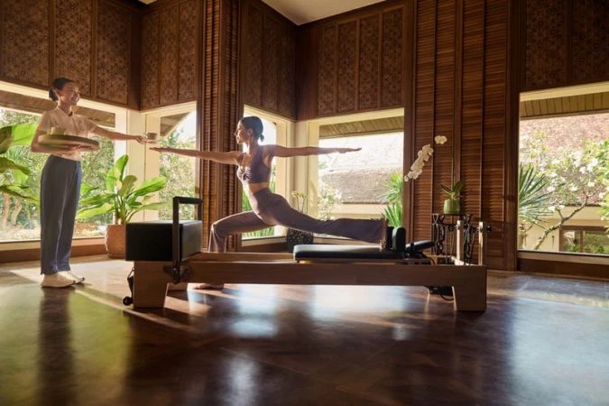 An adult female balances in warrior pose on a Pilates reformer indoors as a server brings her a drink near a window.