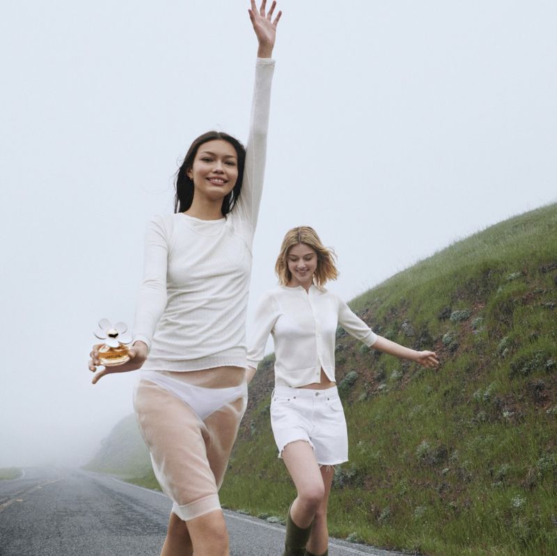 Two young women stroll on a road with grassy hills. One carries a perfume bottle. The setting is an overcast day with muted colors.