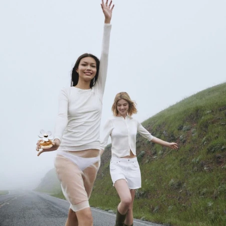 Two young women stroll on a road with grassy hills. One carries a perfume bottle. The setting is an overcast day with muted colors.