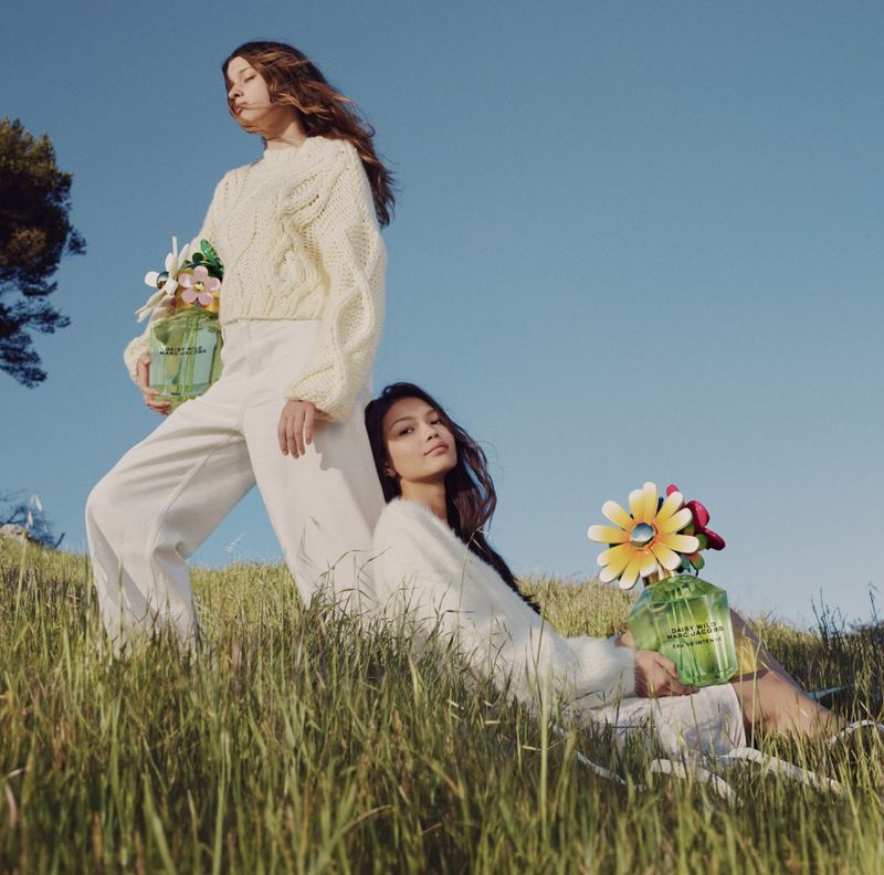 Two women pose in a grassy field with bright sky, holding flower-topped perfume bottles, wearing knitwear, and light pants, low angle.