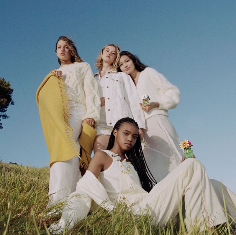 Four women pose in an outdoor field wearing white clothing and holding small vases of flowers against a blue sky backdrop.