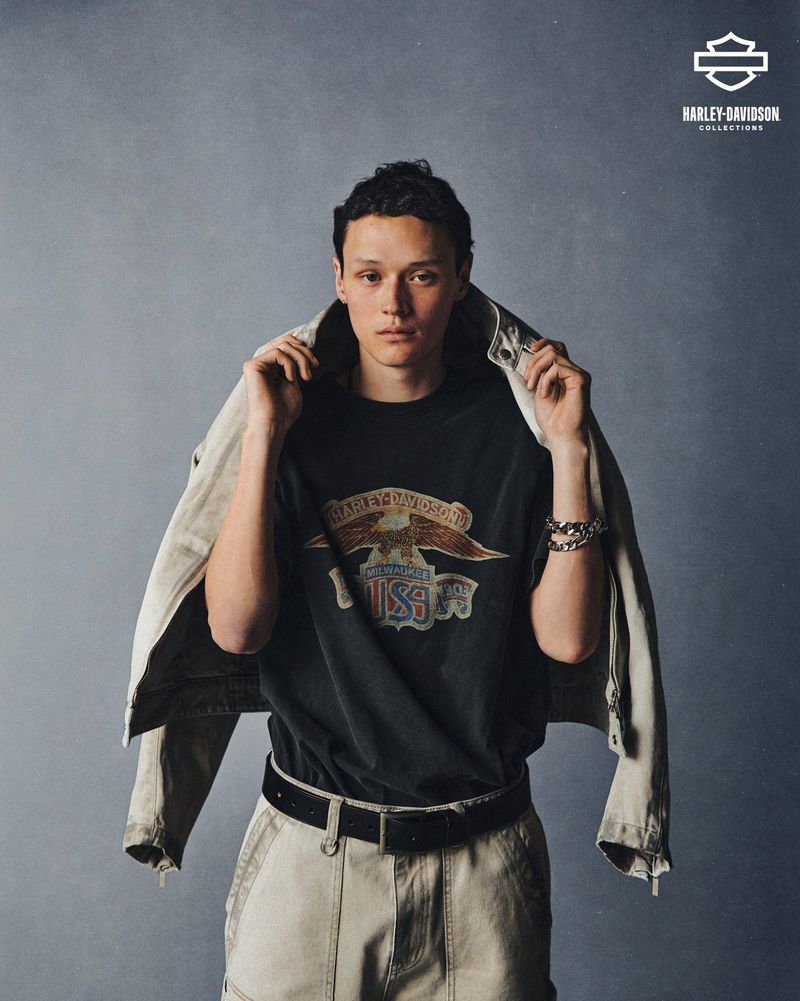 Male fashion model poses wearing a Harley-Davidson graphic t-shirt with a light-colored jacket draped over his shoulders in a studio setting.