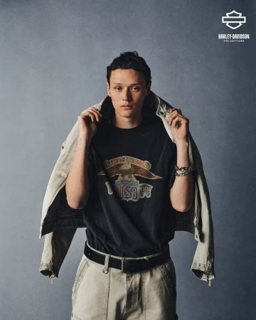Male fashion model poses wearing a Harley-Davidson graphic t-shirt with a light-colored jacket draped over his shoulders in a studio setting.