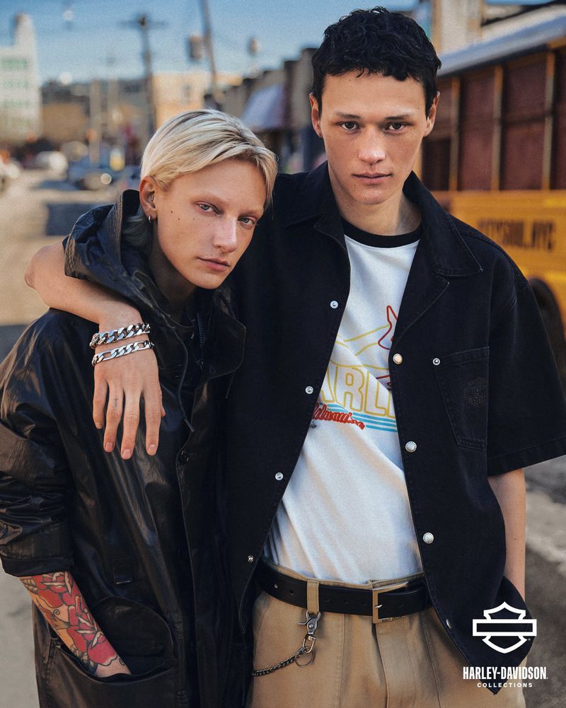 Fashion shot of a blonde adult female and a male model in outdoor urban setting wearing modern casualwear with a motorcycle brand logo.