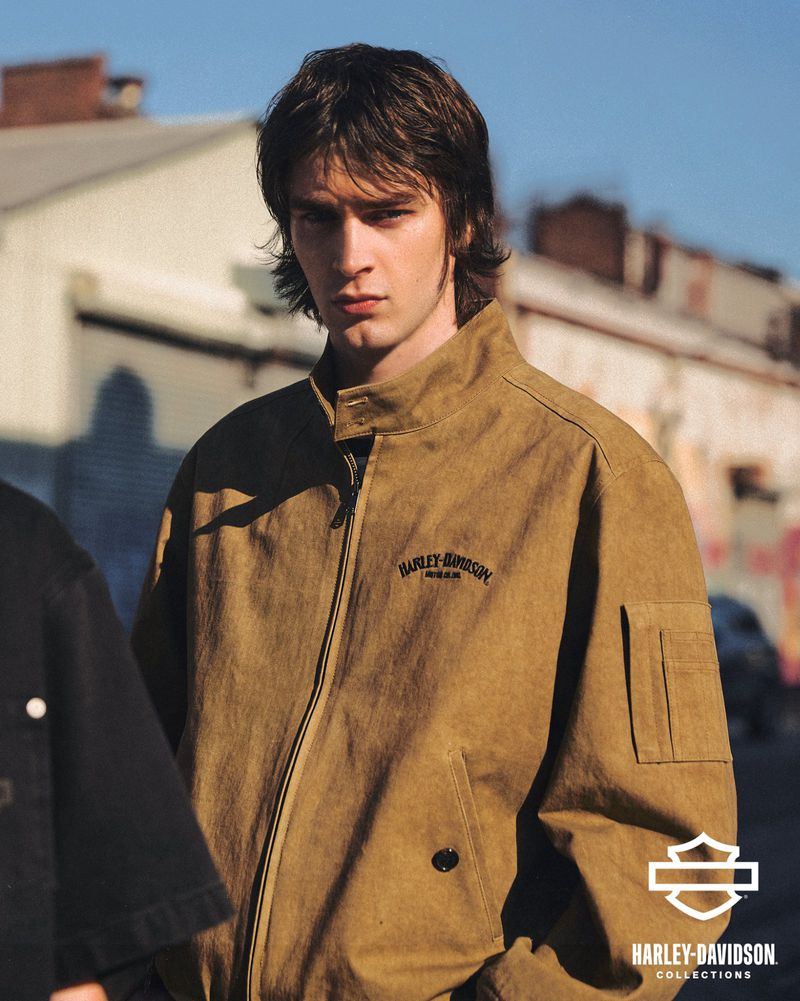 Man in a tan Harley-Davidson bomber jacket poses outdoors. Urban setting with building and street visible in the background during the day.