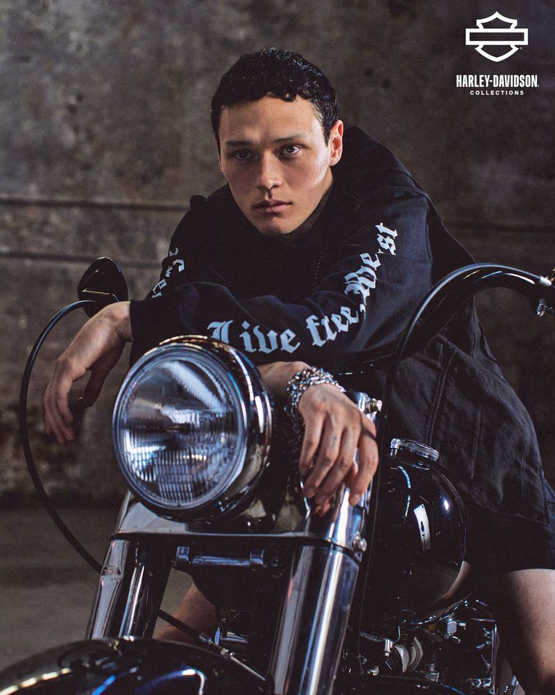 Young male poses with a Harley-Davidson motorcycle indoors. The motorcycle's headlight is prominent, and the subject is wearing a black hooded shirt.