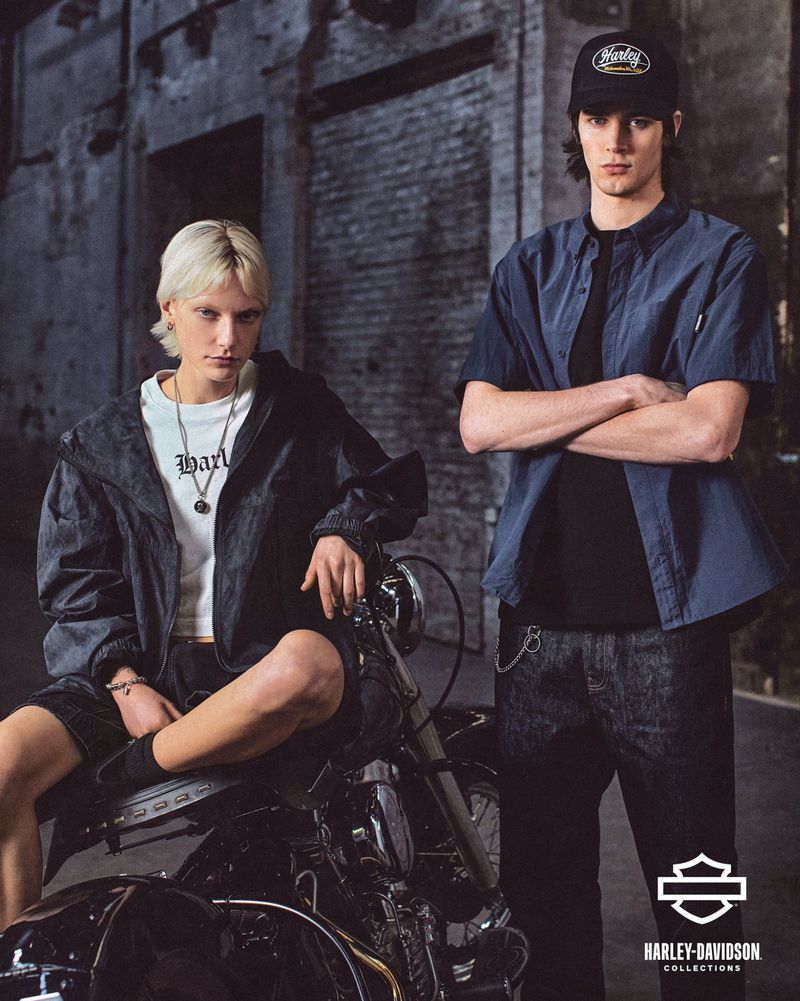 Two young adult models pose with a Harley-Davidson motorcycle on a city street, showcasing fashion apparel from the Harley-Davidson collection.
