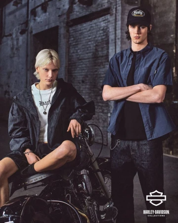 Two young adult models pose with a Harley-Davidson motorcycle on a city street, showcasing fashion apparel from the Harley-Davidson collection.
