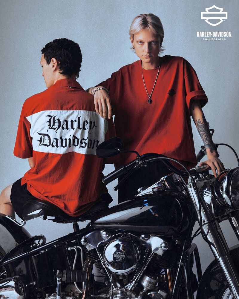 Two people in Harley Davidson apparel next to a motorcycle. One poses; one is looking away. Focus on fashion and branding.