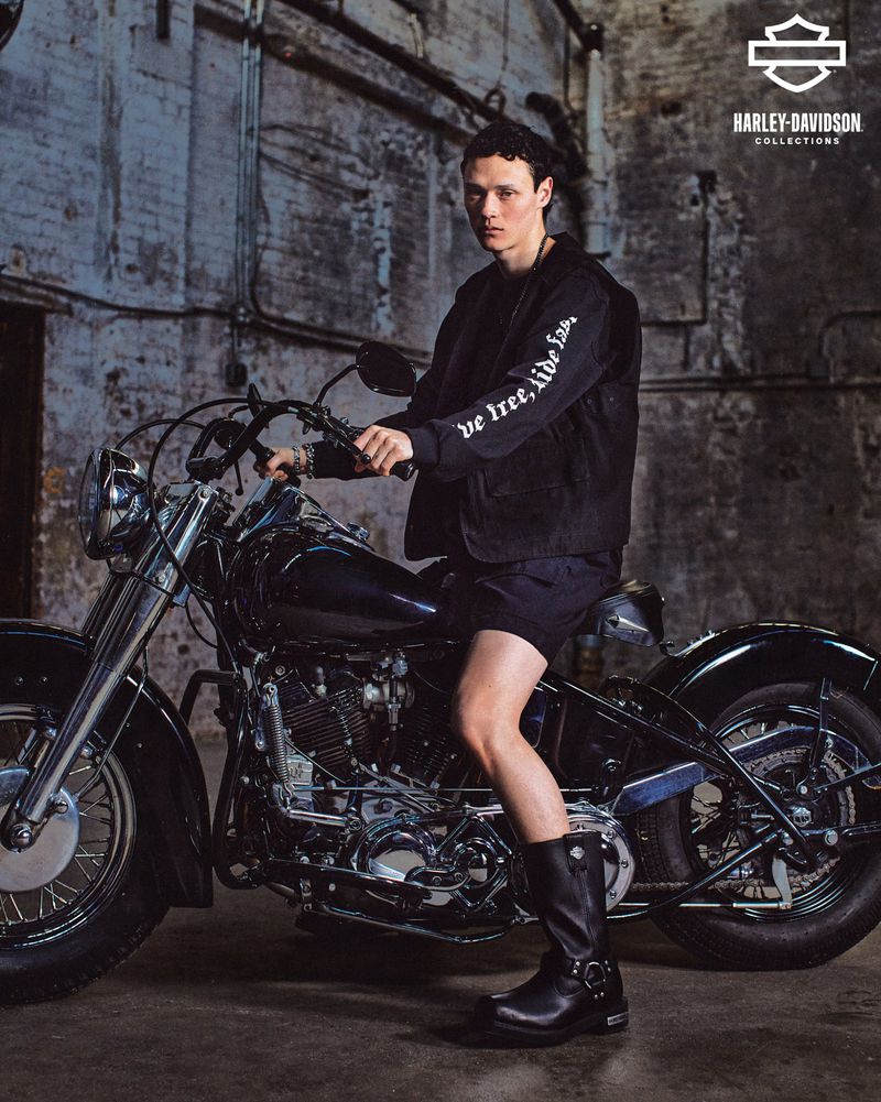 A man in a black jacket sits on a vintage Harley-Davidson motorcycle in an industrial setting. The shot features dramatic lighting and textures.