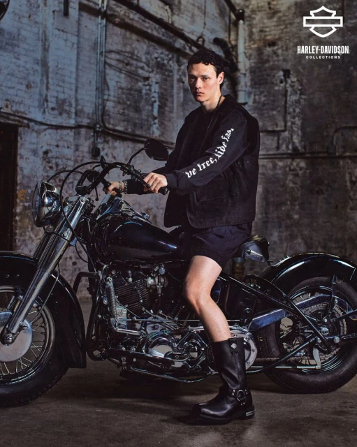 A man in a black jacket sits on a vintage Harley-Davidson motorcycle in an industrial setting. The shot features dramatic lighting and textures.